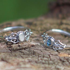 May include: Two silver rings with ornate filigree detailing. One ring features a white oval stone, the other a blue and green abalone shell. The rings are displayed on a textured, brown surface.