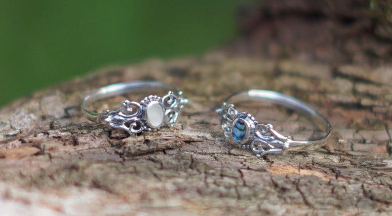 May include: Two silver rings with ornate filigree detailing. One ring features a white oval stone, the other a blue and green abalone shell. The rings are displayed on a textured, brown surface.