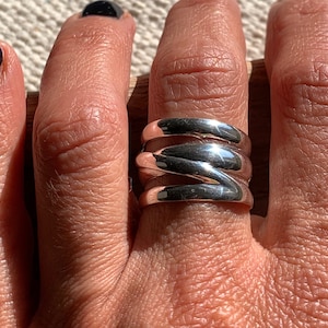 Pu&ograve; includere: Un anello d'argento con un design ondulato a pi&ugrave; strati. L'anello &egrave; indossato su un dito.