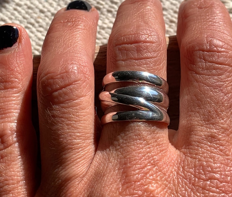 Pu&ograve; includere: Un anello d'argento con un design ondulato a pi&ugrave; strati. L'anello &egrave; indossato su un dito.