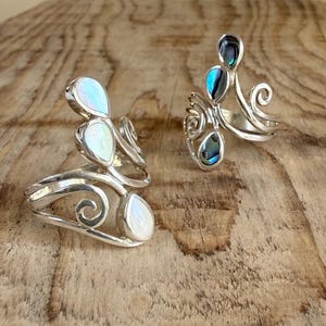 Può includere: Due anelli in argento con fasce metalliche a spirale e inserti iridescenti a forma di goccia. L'anello a sinistra presenta pietre bianche madreperlacee, mentre quello a destra ha accenti in conchiglia di abalone blu-verde.