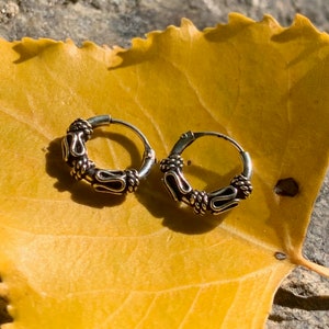 Peut inclure: Deux boucles d'oreilles créoles en argent avec un design détaillé et complexe. Les boucles d'oreilles sont sur une feuille jaune.