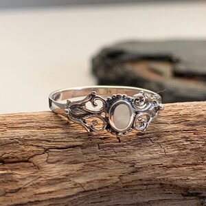 May include: A silver ring with an oval, off-white stone set in an ornate, filigree design. The ring is displayed on a piece of weathered wood, showcasing its intricate details and vintage style.