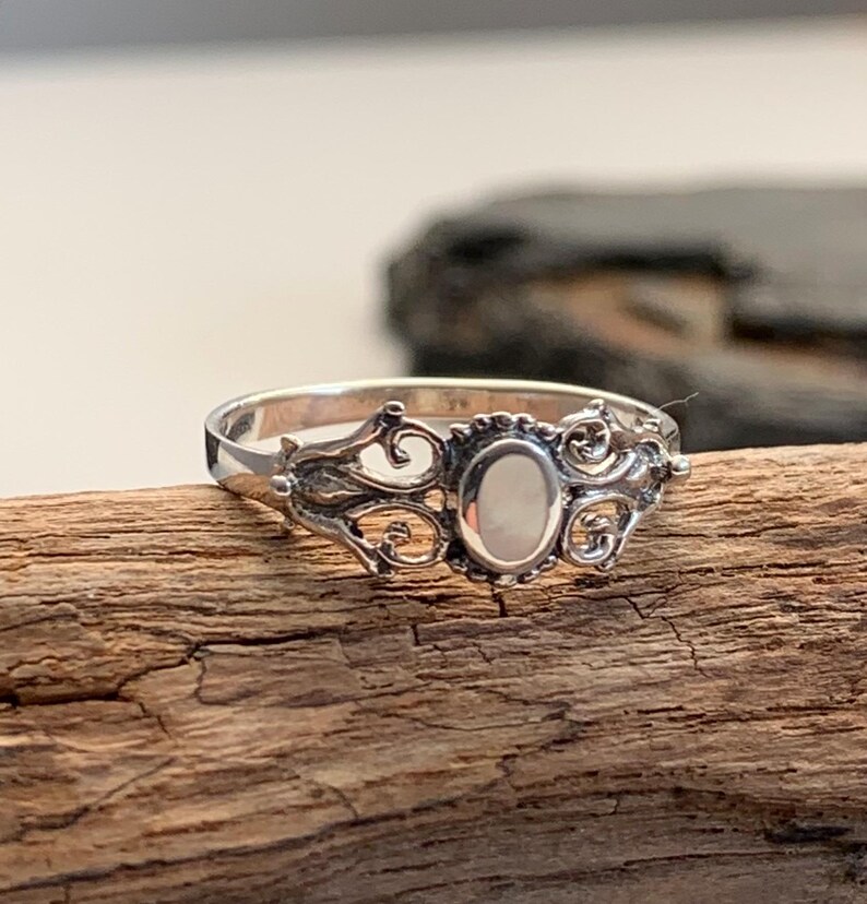 May include: A silver ring with an oval, off-white stone set in an ornate, filigree design. The ring is displayed on a piece of weathered wood, showcasing its intricate details and vintage style.