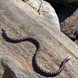 K&ouml;nnte beinhalten: Ein silbernes Armband mit Perlen und einem Verschluss. Das Armband liegt auf einem grauen Stein.