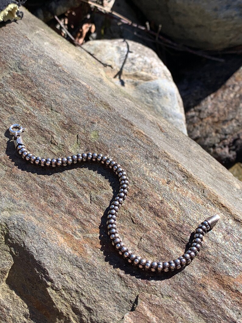 K&ouml;nnte beinhalten: Ein silbernes Armband mit Perlen und einem Verschluss. Das Armband liegt auf einem grauen Stein.