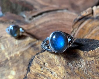 Op de afbeelding: Twee zilveren ringen met ovale, blauwe edelstenen. De ringen zijn geplaatst op een houten oppervlak. De edelstenen hebben een blauwe iridescentie. De ringen hebben een decoratieve band met kleine, ronde accenten.