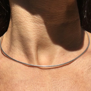 Op de afbeelding: Een zilveren chokerketting met een dun, glad en afgerond ontwerp. De ketting wordt gedragen door een persoon met een lichte huidskleur.