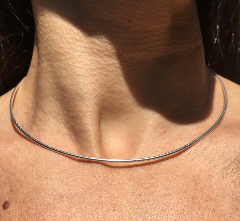 Op de afbeelding: Een zilveren chokerketting met een dun, glad en afgerond ontwerp. De ketting wordt gedragen door een persoon met een lichte huidskleur.
