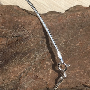Op de afbeelding: Een zilveren slangenketting met een karabijn sluiting. De ketting ligt op een stuk hout.