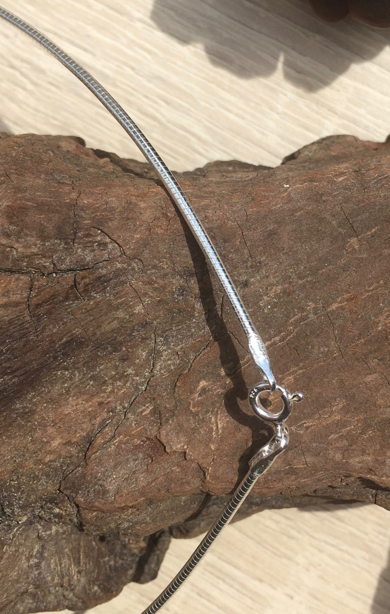 Op de afbeelding: Een zilveren slangenketting met een karabijn sluiting. De ketting ligt op een stuk hout.