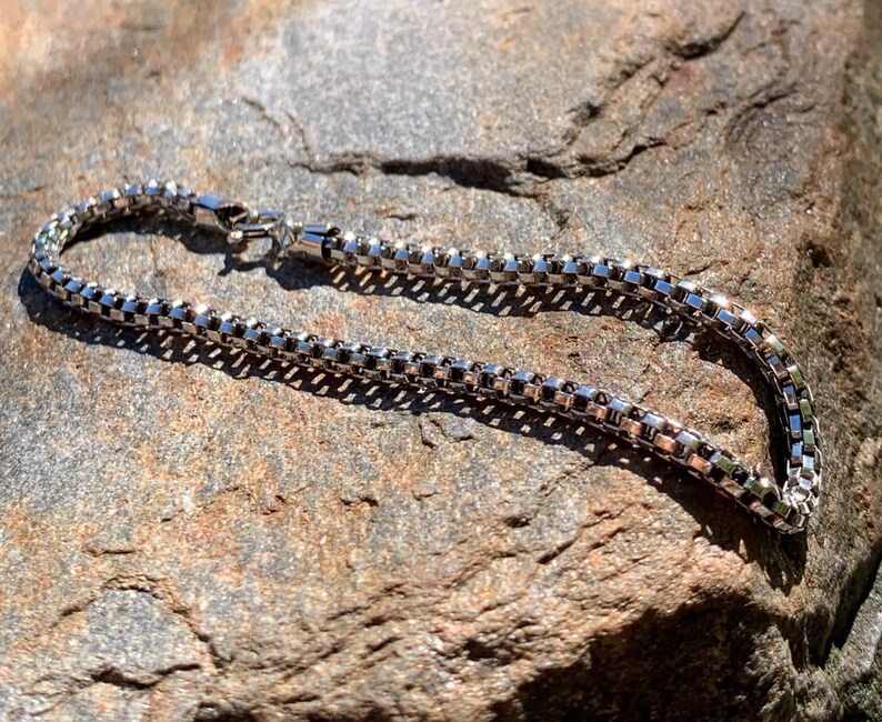 May include: A silver chain necklace with a box clasp. The chain is made of small, square links.