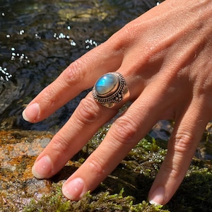 Op de afbeelding: Een zilveren ring met een grote, ovale, iriserende blauwe edelsteen. De ring heeft een gedetailleerde, getextureerde band en wordt om een vinger gedragen. De achtergrond toont water en groen.
