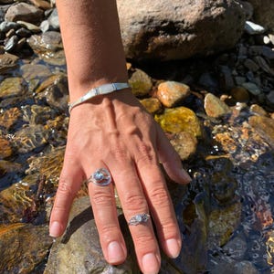 May include: A silver bracelet and two rings with iridescent stones. The bracelet has rectangular mother-of-pearl inlays. One ring features a circular design with a blue-green stone, and the other has a floral filigree setting with a similar stone.