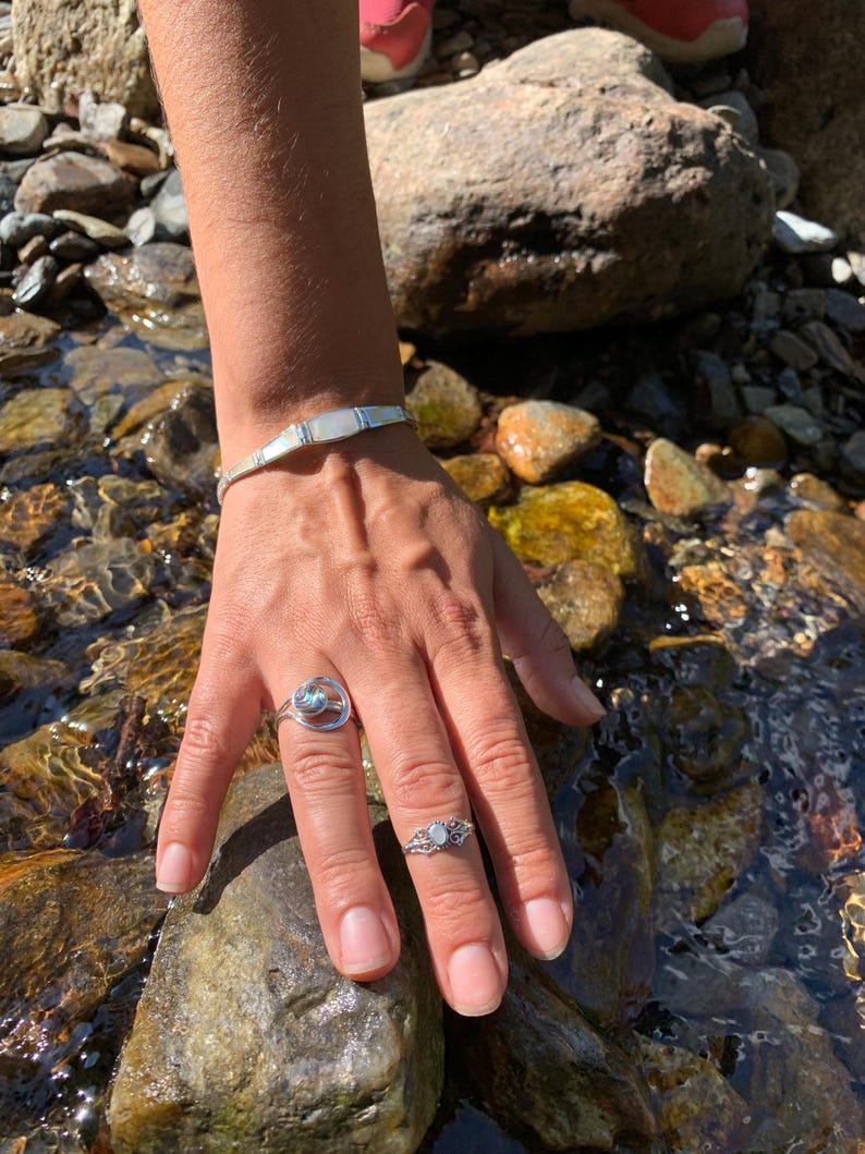 May include: A silver bracelet and two rings with iridescent stones. The bracelet has rectangular mother-of-pearl inlays. One ring features a circular design with a blue-green stone, and the other has a floral filigree setting with a similar stone.
