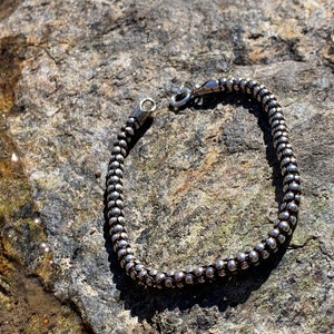 K&ouml;nnte beinhalten: Ein silbernes Armband mit Perlen und einem Verschluss. Das Armband liegt auf einem grauen Stein.
