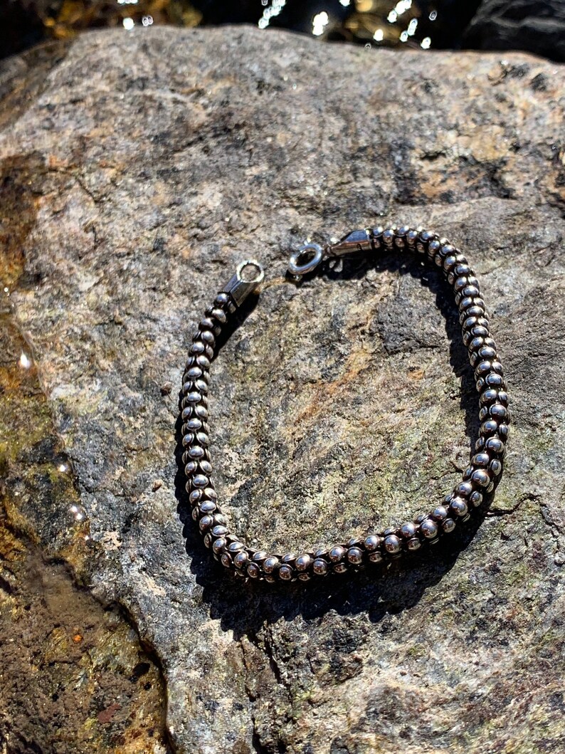 K&ouml;nnte beinhalten: Ein silbernes Armband mit Perlen und einem Verschluss. Das Armband liegt auf einem grauen Stein.