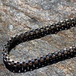 May include: A silver chain necklace with a box chain design. The chain is lying on a textured surface.