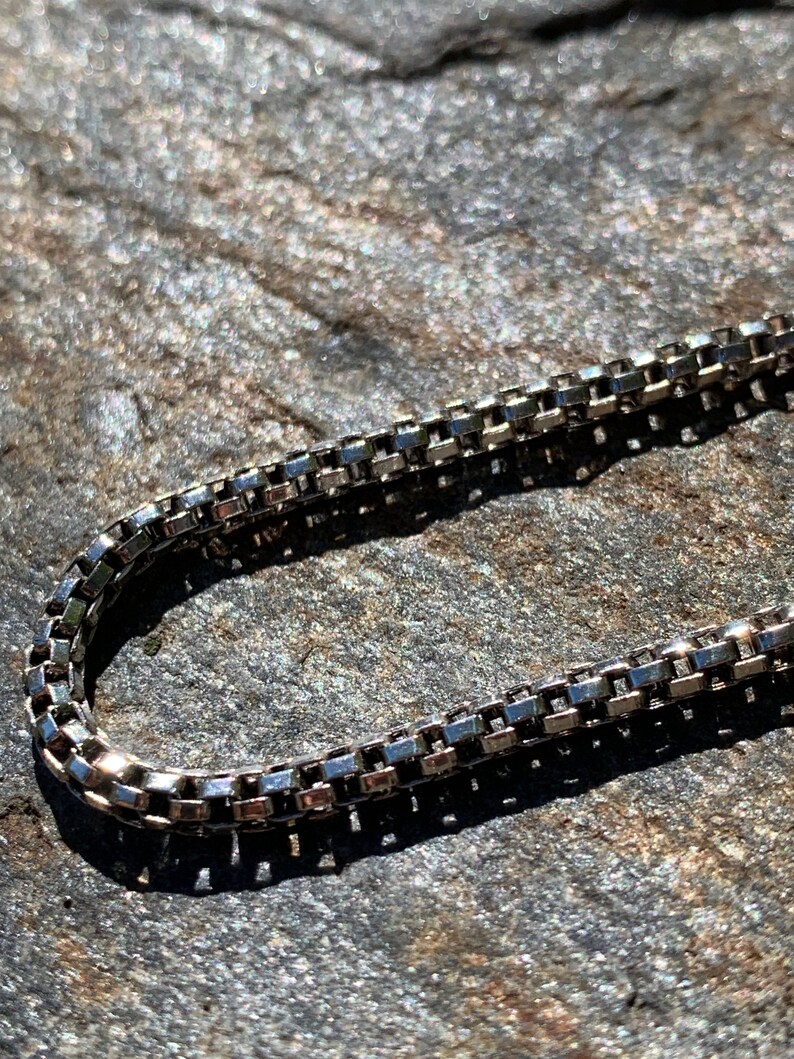May include: A silver chain necklace with a box chain design. The chain is lying on a textured surface.