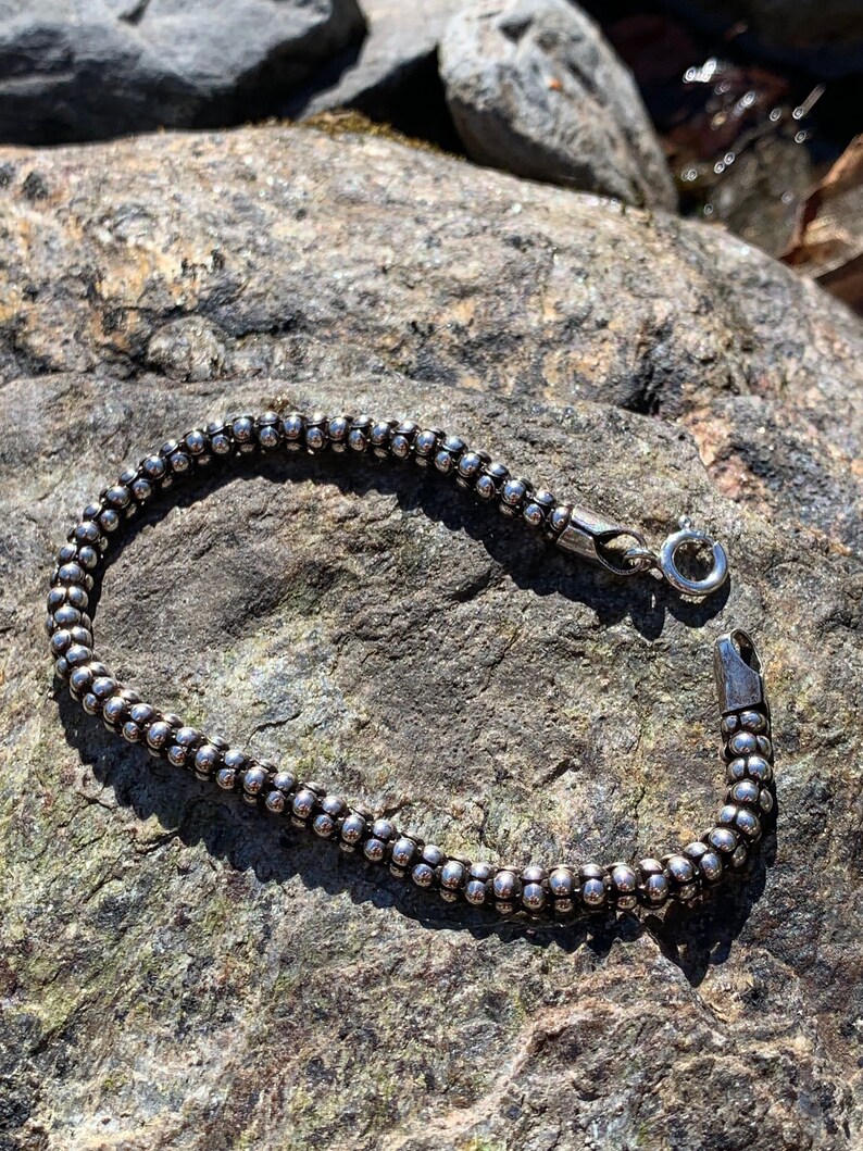 K&ouml;nnte beinhalten: Ein silbernes Armband mit Perlen und einem Karabinerverschluss. Das Armband liegt auf einer rauen, grauen Steinoberfl&auml;che.