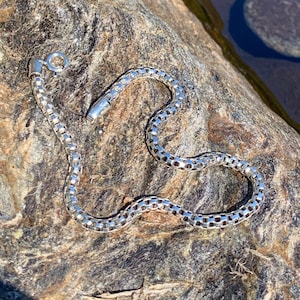 May include: A silver chain necklace with a box chain design. The chain is lying on a textured rock surface.