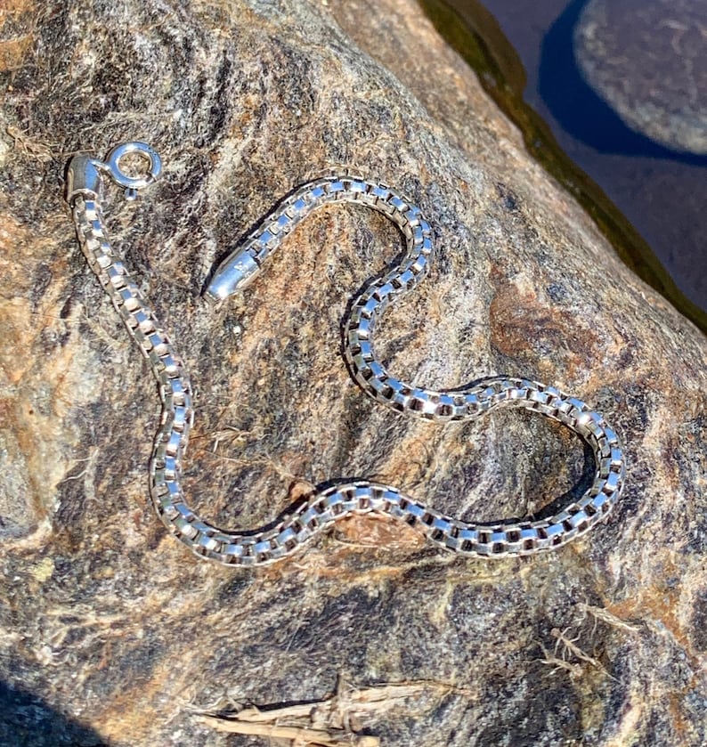 May include: A silver chain necklace with a box chain design. The chain is lying on a textured rock surface.