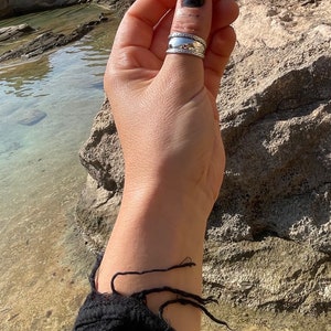May include: A silver ring with a textured band and a smooth, rounded top. The ring is worn on the thumb of a hand.