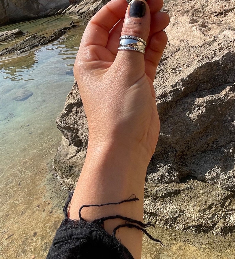 May include: A silver ring with a textured band and a smooth, rounded top. The ring is worn on the thumb of a hand.