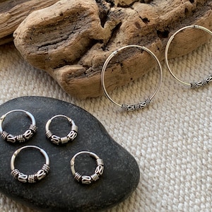 Peut inclure: Quatre boucles d'oreilles créoles en argent avec des perles décoratives. Les boucles d'oreilles sont disposées sur une pierre grise et un morceau de bois flotté.