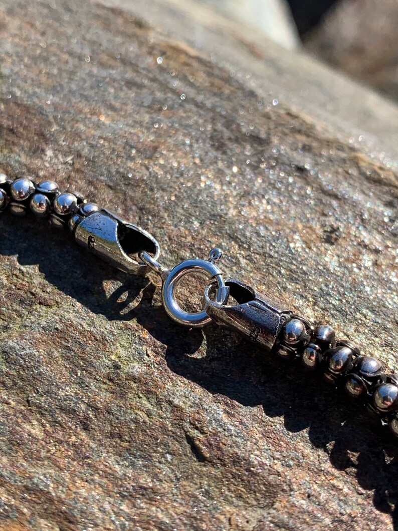 K&ouml;nnte beinhalten: Nahaufnahme einer silbernen Kettenkette mit einem Karabinerverschluss. Die Kette besteht aus kleinen, runden Perlen.