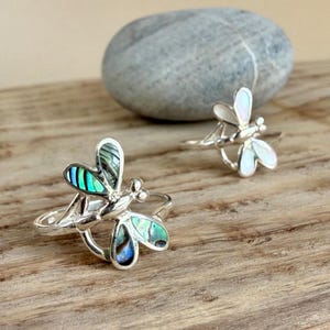 Könnte beinhalten: Zwei silberne Ringe in Libellenform. Der vordere Ring hat Flügel, die mit schillerndem grün-blauem Abalone-Muschelmaterial eingelegt sind. Der hintere Ring hat Flügel aus weißem Perlmutt. Beide Ringe verfügen über eine polierte silberne Ringschiene.