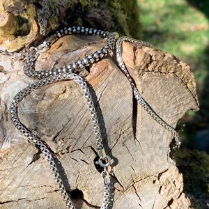 May include: Two silver chains, one box chain and one curb chain, are displayed on a piece of weathered wood.