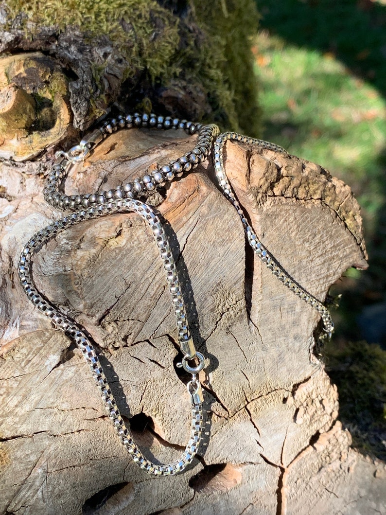 May include: Two silver chains, one box chain and one curb chain, are displayed on a piece of weathered wood.