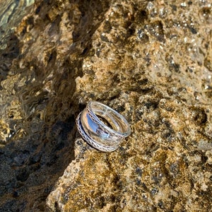 May include: A silver ring with a twisted design, resting on a textured rock surface.