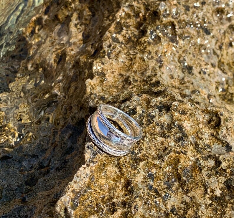 May include: A silver ring with a twisted design, resting on a textured rock surface.