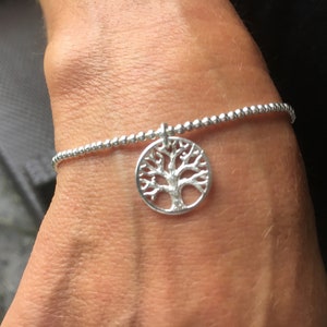 May include: Silver beaded bracelet featuring a tree of life charm. The charm is a circular, detailed cutout of a tree. The bracelet is worn on a wrist.