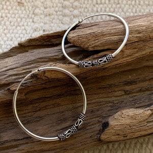 May include: A pair of silver hoop earrings with a decorative design. The hoops are thick and have a smooth finish. The design is a series of small, intricate shapes that are repeated around the hoop.