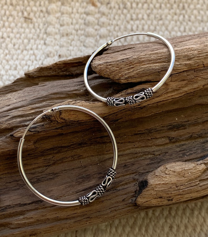 May include: A pair of silver hoop earrings with a decorative design. The hoops are thick and have a smooth finish. The design is a series of small, intricate shapes that are repeated around the hoop.