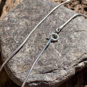 Op de afbeelding: Een close-up van een zilveren slangencolier met een karabijnsluiting. De ketting wordt getoond op een grijze, getextureerde steen. De ketting is dun en flexibel en reflecteert licht.