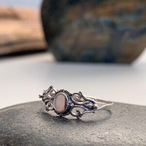 May include: A silver ring with an oval, light-colored stone at the center. The ring features an ornate, swirling design around the stone and along the band. The ring is set on a dark gray surface.