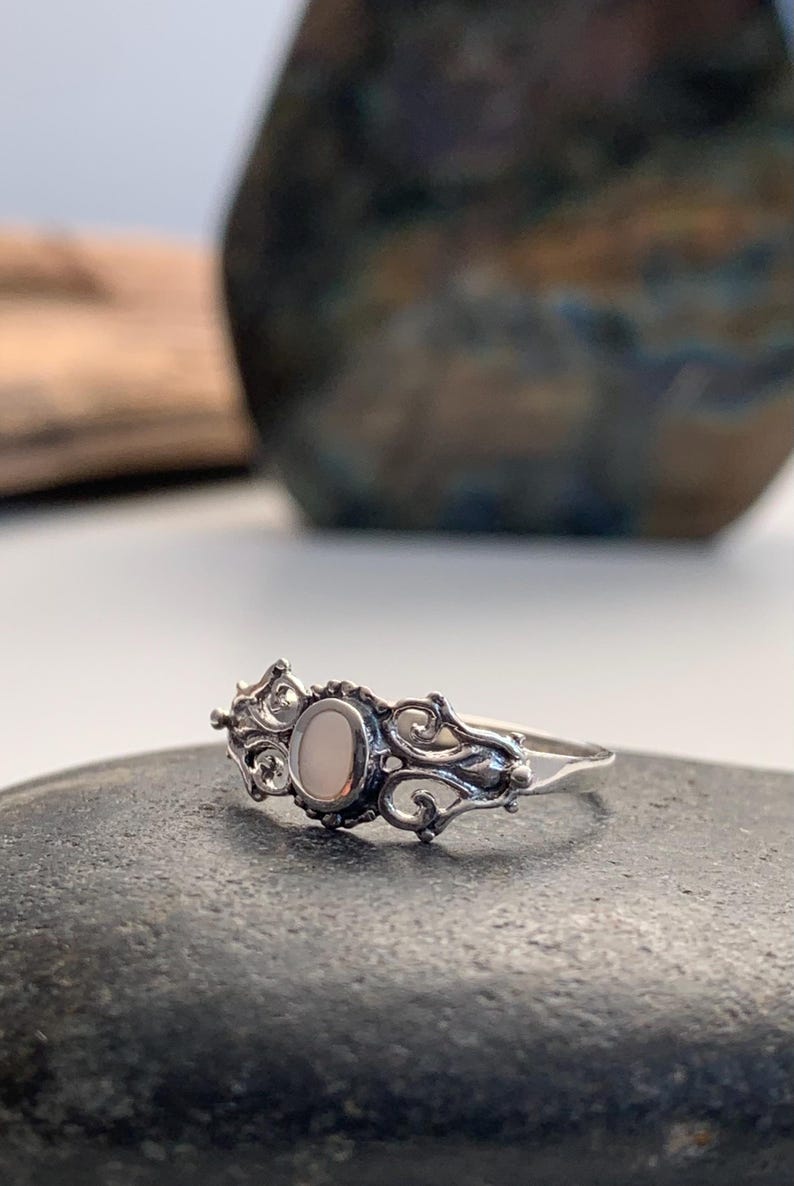 May include: A silver ring with an oval, light-colored stone at the center. The ring features an ornate, swirling design around the stone and along the band. The ring is set on a dark gray surface.