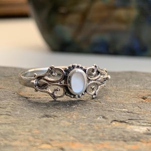 May include: A silver ring with an oval, white gemstone center. The ring features ornate, swirling filigree designs on either side of the gemstone. The ring is set on a gray, textured surface, with a blurred background.