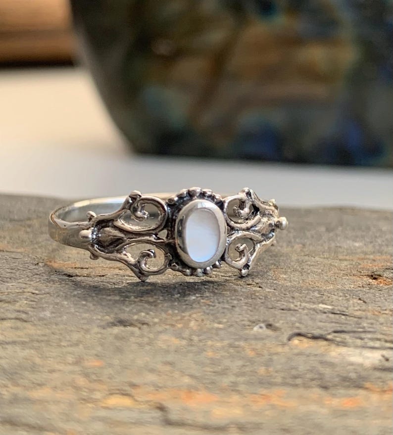 May include: A silver ring with an oval, white gemstone center. The ring features ornate, swirling filigree designs on either side of the gemstone. The ring is set on a gray, textured surface, with a blurred background.
