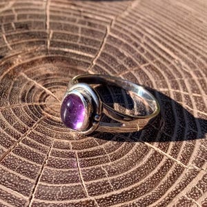 Pode incluir: Um anel de prata com uma pedra preciosa ametista roxa de formato oval e lisa, engastada numa moldura de metal polido. A banda tem um design dividido que se estreita para a parte de trás, exibindo um acabamento metálico refletor.