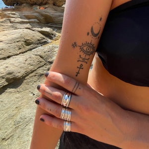 May include: A person's hand wearing a stack of four silver rings. The rings are all different sizes and have a textured design. The person's arm has a tattoo of a crescent moon, stars, an eye, and a cross.