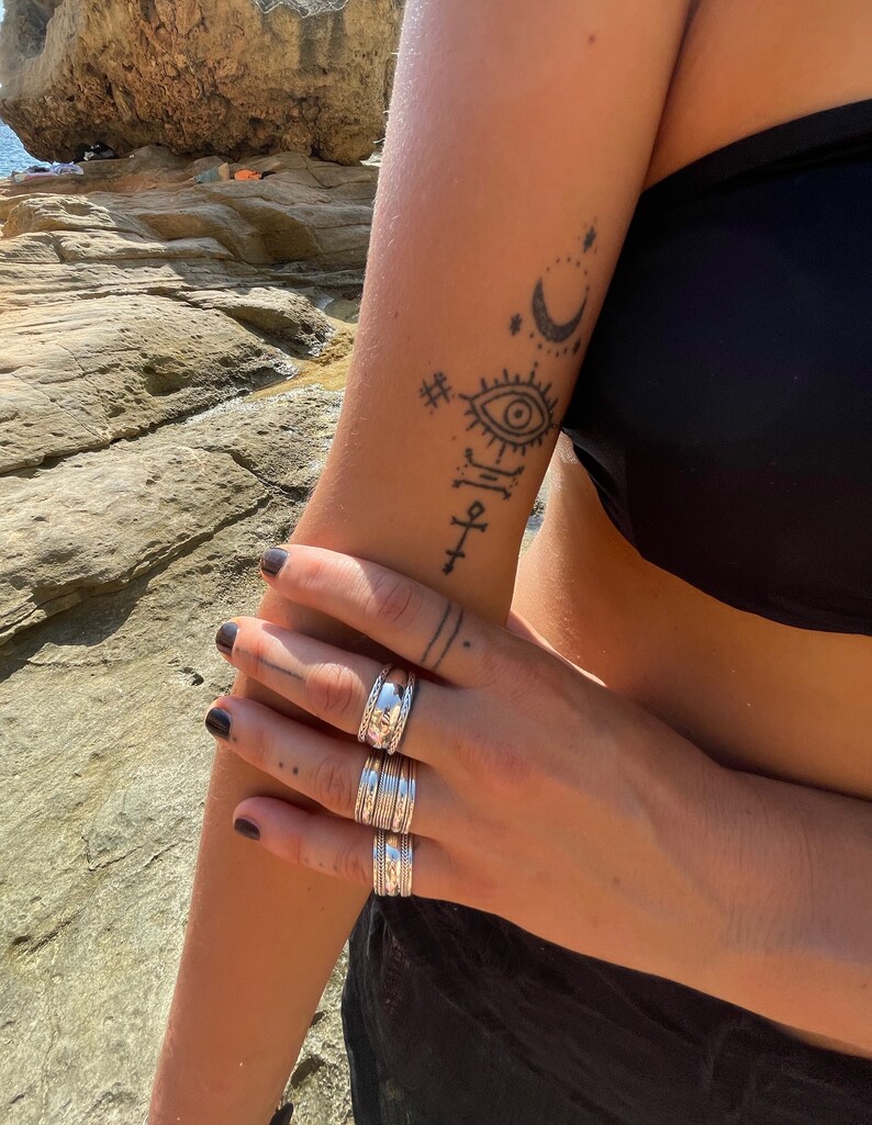 May include: A person's hand wearing a stack of four silver rings. The rings are all different sizes and have a textured design. The person's arm has a tattoo of a crescent moon, stars, an eye, and a cross.