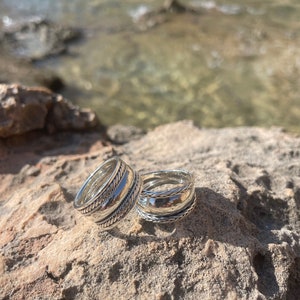 May include: Two silver rings with intricate designs, stacked on top of each other, resting on a rocky surface.