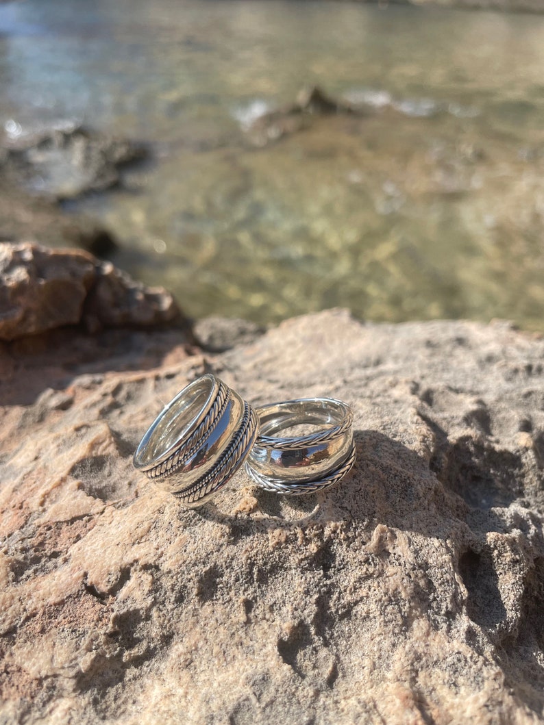 May include: Two silver rings with intricate designs, stacked on top of each other, resting on a rocky surface.