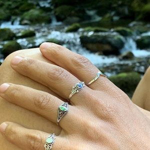 May include: Close-up of a hand wearing four silver rings. The rings feature various designs, including a curved band, an oval-shaped setting with a green stone, and two rings with ornate, filigree-style settings and colored stones.