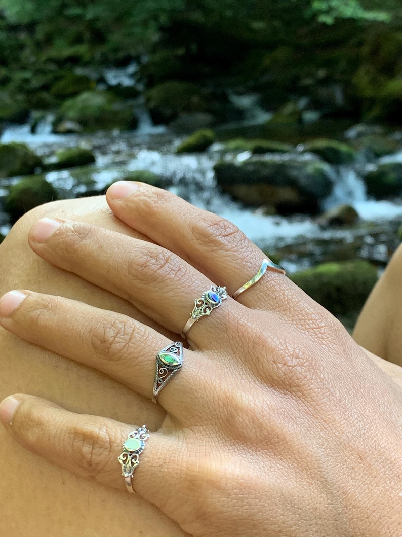 May include: Close-up of a hand wearing four silver rings. The rings feature various designs, including a curved band, an oval-shaped setting with a green stone, and two rings with ornate, filigree-style settings and colored stones.